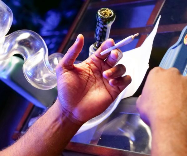 "A person holding a lit joint and a bong with cannabis, preparing to smoke in a dimly lit environment with a blue glow, highlighting the disadvantages of drug use."