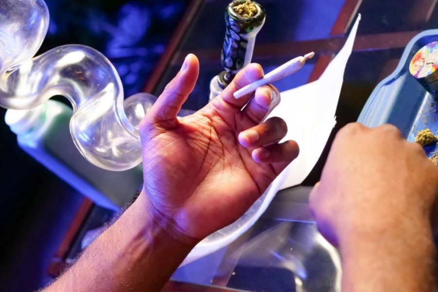"A person holding a lit joint and a bong with cannabis, preparing to smoke in a dimly lit environment with a blue glow, highlighting the disadvantages of drug use."